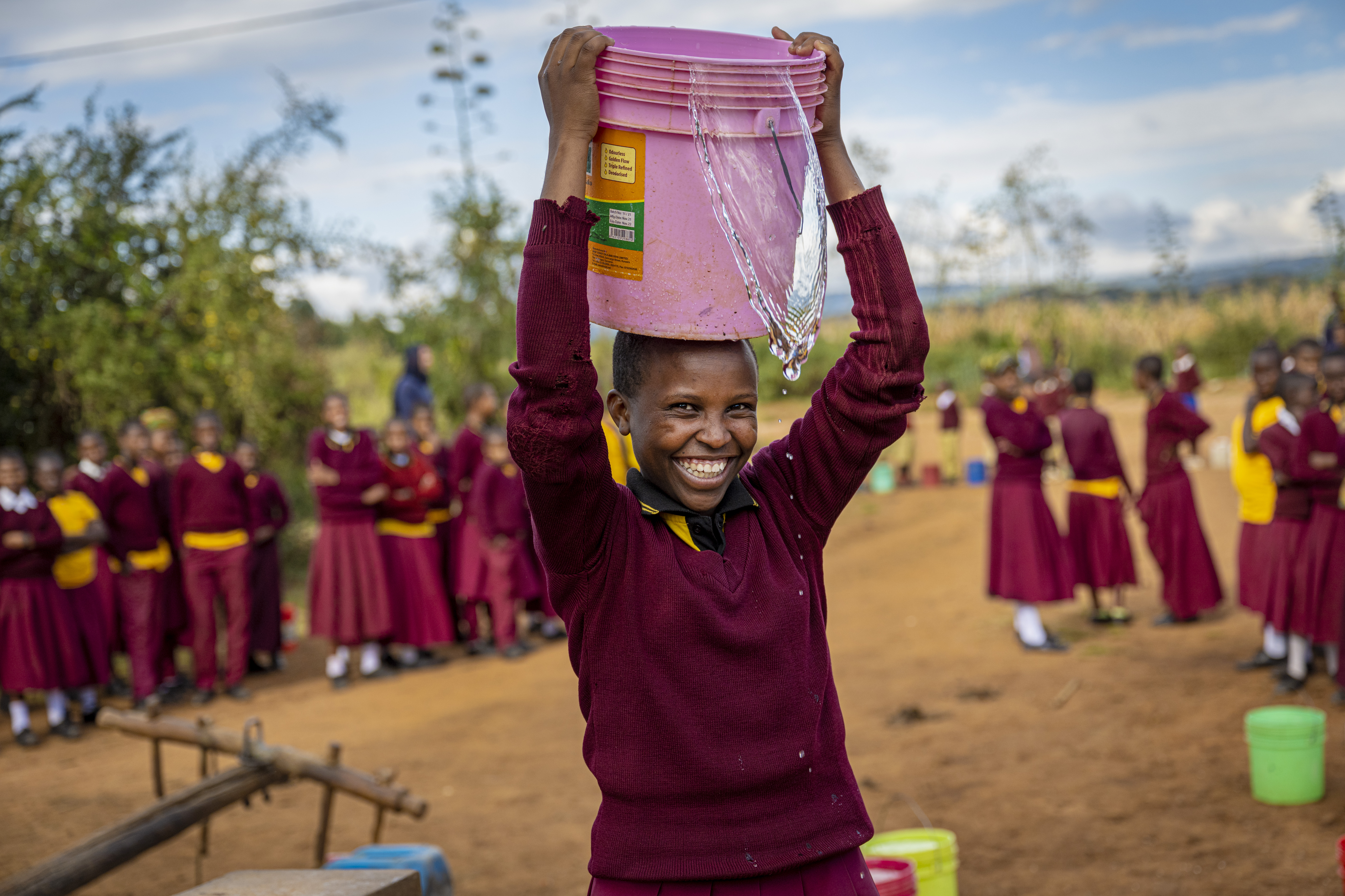 15 år gamle Deodata fra Tanzania har på skoleuniform med mørk rød genser og skjørt. Hun holder ei rosa bøtte med vann på hodet. Bak henne står klassekameratene hennes. I bakgrunnen er det busker og trær og blå himmel.